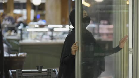 Woman-in-hijab-and-protective-mask-doing-shopping,-takes-product-from-the-freezer-shelf