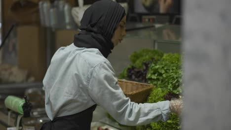 Woman-in-scarf-refill-the-fresh-greens-on-the-shelf-at-the-supermarket,-side-view
