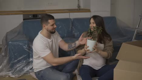 Smiling-couple-sitting-on-the-floor-in-the-living-room,-unpacking-plant-from-the-box