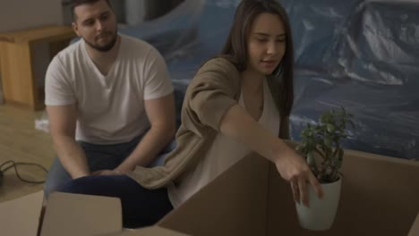 Cheerful-couple-sitting-on-the-floor-in-the-living-room,-unpacking-plant-from-the-box