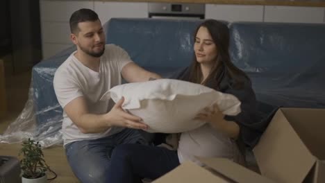 Married-couple-sitting-on-the-floor-in-the-living-room,-unpacking-pillow-from-the-box