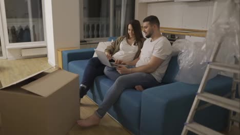 Eine-Schwangere-Frau-Und-Ein-Positiver-Mann-Sitzen-Mit-Einem-Laptop-Auf-Dem-Sofa-In-Einem-Neuen-Wohnzimmer