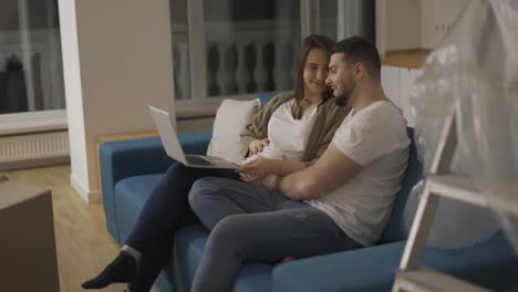 Lovely-pregnant-woman-and-a-positive-man-are-using-a-laptop-sitting-on-the-sofa-in-a-new-living-room