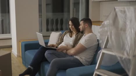 Lovely-pregnant-woman-and-a-man-are-using-a-laptop-sitting-on-the-sofa-in-a-new-living-room