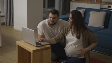 Schöne-Schwangere-Frau-Und-Mann-Surfen-Im-Internet-Und-Sitzen-Mit-Einem-Laptop-Auf-Dem-Boden