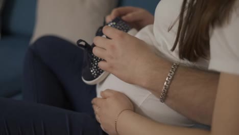 Beautiful-pregnant-woman-and-her-husband-holding-baby-shoes-on-belly,-close-up