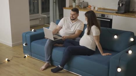 Hübsches-Schwangeres-Paar,-Das-Einen-Laptop-Benutzt-Und-Auf-Dem-Sofa-In-Einem-Neuen-Wohnzimmer-Mit-Girlanden-Sitzt