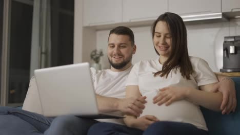 Schöne-Schwangere-Frau-Und-Mann-Surfen-Im-Internet-Und-Sitzen-Auf-Der-Couch-Mit-Laptop