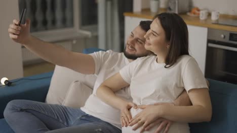 Happy-man-and-his-pregnant-wife-taking-selfie-by-smartphone-at-home