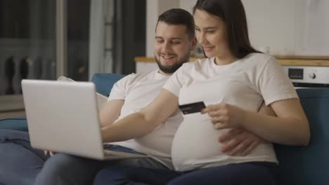 Couple-expecting-baby,-sitting-on-sofa-at-home-and-shopping-online-using-laptop