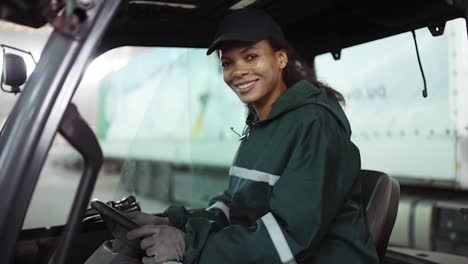Potrait-of-an-African-American-woman-in-a-special-green-uniform-sitting-behind-the-wheel-of-a-truck-in-a-waste-processing-plant.-Processing-of-raw-materials,-recycling.-Pollution-control