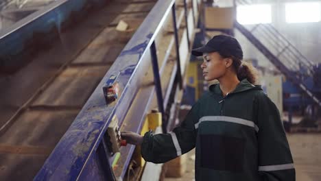 Young-african-american-woman-checking-conveyor-belt-with-push-buttons-at-waste-recycling-plant.-Pollution-control