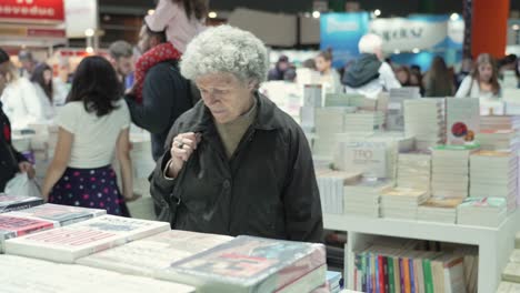 Elderly-woman-walks-past-and-browses-looking-at-stacks-of-books-at-fair