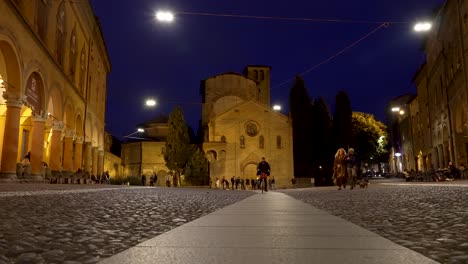A-low-angle-shot-of-the-Basilica-of-Santo-Stefano-illuminated-by-lights-on-a-beautiful-evening,-as-a-couple-walk-past-enjoying-a-relaxing-walk-through-the-city-at-night,-Bologna,-Italy