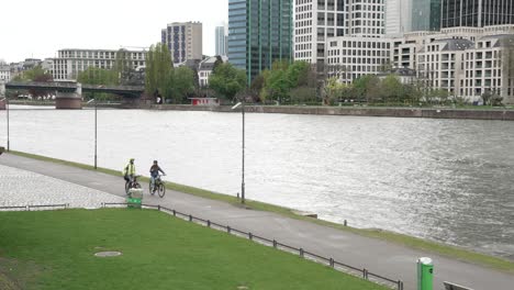 Pair-Of-Cyclists-Riding-Along-River-Main-In-Downtown-Frankfurt