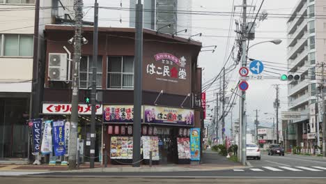 Straßenecke-In-Der-Innenstadt-Von-Hakodate-Mit-Vorbeifahrenden-Geschäften-Mit-Bunten-Ladenfrontautos