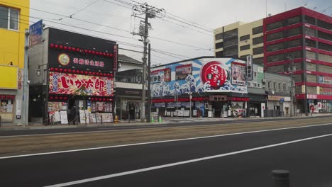Straße-In-Der-Innenstadt-Von-Hakodate-Mit-Geschäften-Mit-Farbenfroher-Ladenfront-Und-Vorbeifahrenden-Blauen-Lokalen-Taxis
