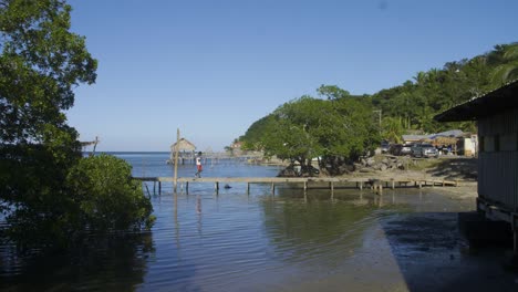 Punta-Gorda-beach-at-Roatan,-Honduras-during-the-daytime-as-person-walks-across-a-wooden-pier-towards-the-road