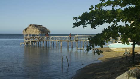 Punta-Gorda-beach-at-Roatan,-Honduras-during-the-daytime-as-person-walks-across-a-rustic-wooden-pier-towards-the-beach