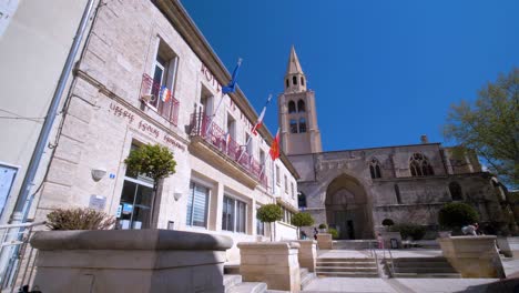 Sunny-Day-at-Montagnac-Town-Hall-Square:-French,-European,-and-Occitanie-Flags-Fluttering,-with-the-Village-Church-in-the-Background