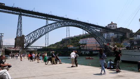 Panning-shot-of-tourist-in-city-port-of-Porto-with-famous-Ponte-Dom-Luís-I-during-sunny-day---Slow-motion-wide-shot-on-promenade