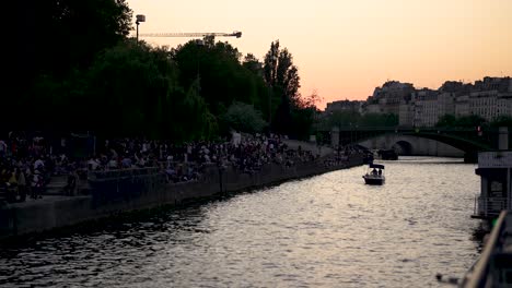 People-on-the-shore-of-the-Seine-river-at-sunset-seen-from-river-boat,-Left-view-backward-shot