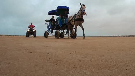 Harnessed-horse-pulling-carriage-with-tourists-along-Tunisia-arid-desert-with-quad-driving-behind