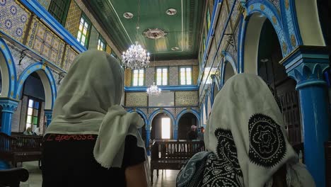Back-view-of-two-tourist-women-with-headscarf-sitting-inside-El-Ghriba-Jewish-synagogue-of-Djerba-in-Tunisia