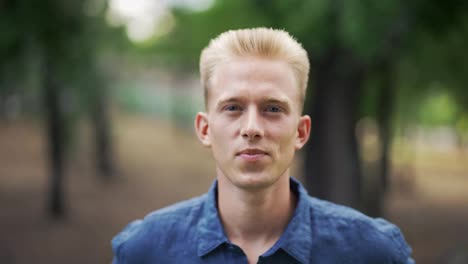Close-up-portrait-young-happy-blond-man-standing-in-nature-between-park-trees-relaxes,-breathes-fresh-air.-Male-enjoys-a-life-of