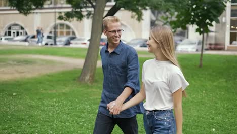 A-young-positive-couple-walks-holding-hands,-meets-in-a-summer-city-park.-The-girl-and-the-boy-are-blonde.-Happy-walk-in-the-park