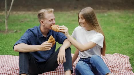 A-girl-and-a-guy-feed-each-other-a-croissant-at-a-picnic-as-a-couple.-romantic-date-happiness