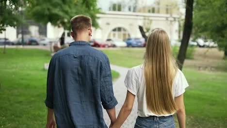 A-young-positive-couple-walks-holding-hands,-meets-in-a-summer-city-park.-The-girl-and-the-boy-are-blonde.-Happy-walk-in-the-park.-Back-view