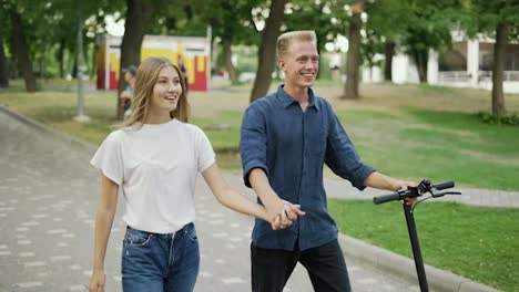 The-guy-and-the-girl-are-walking-in-the-park,-talking.-The-guy-is-holding-a-scooter-in-his-hand.-Happy-walk.-romantic-date