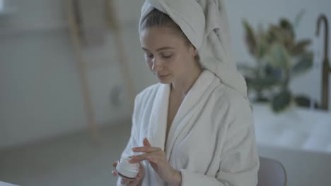 Young-woman-in-white-bathrobe-and-with-towel-on-head-applies-moisturizer-cream-on-her-face