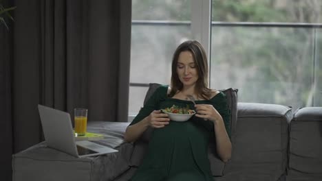 Pregnant-woman-sitting-on-the-sofa-at-home-using-laptop-and-eats-salad