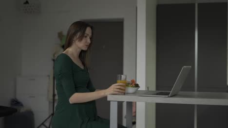 Pregnant-woman-sitting-on-the-kitchen-eats-salad-while-having-a-video-call-on-laptop