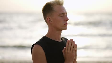 Portrait-of-a-young-man-meditating-against-the-backdrop-of-the-waves-of-the-sea-during-dawn.-Close-up.-Hands-near-the-chest