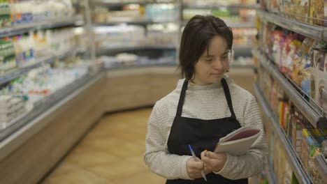Mujer-Con-Síndrome-De-Down-Inspeccionando-Estantes-Con-Productos-En-Una-Tienda-De-Comestibles-Usando-Un-Cuaderno