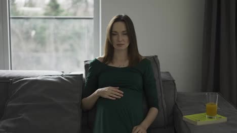 Portrait-of-beautiful-pregnant-woman-sitting-on-sofa-and-gently-touching-her-belly