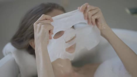 Young-domestic-woman-posing-apply-tissue-face-mask-for-skin-care-while-lying-in-the-bath