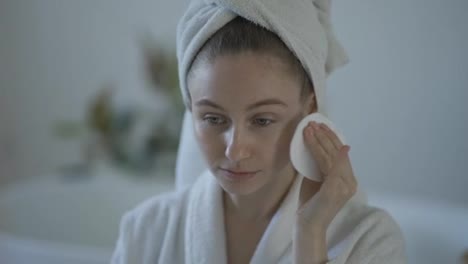 Portrait-of-beautiful-girl-in-bathrobe-and-with-towel-on-her-head-cleaning-her-face-with-sponge