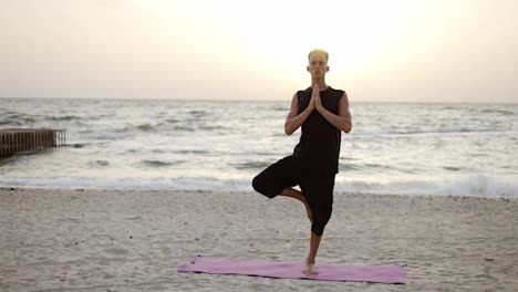Portrait-of-a-young-man-who-practices-yoga-on-a-sports-mat,-standing-on-one-leg-at-dawn.-Performing-a-specific-exercise