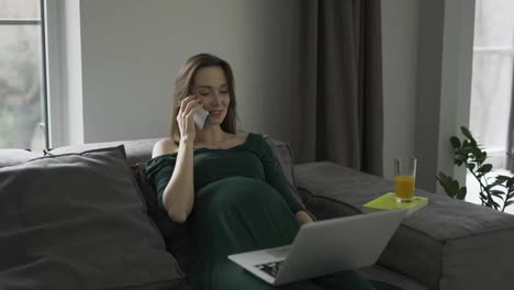 Pregnant-businesswoman-talking-mobile-phone-at-home-office-while-working-on-laptop,-slow-motion