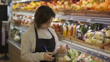 Girl-with-Down-syndrome-inspecting-shelves-with-fresh-fruits-in-a-grocery-store-using-notebook