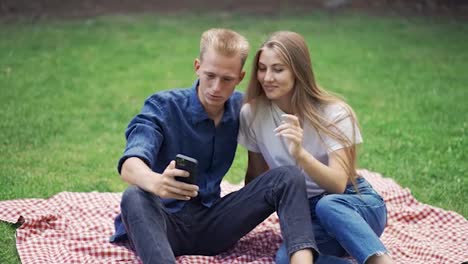 A-guy-and-a-girl-on-a-picnic-take-a-selfie.-People-sit-on-a-mat-in-the-park.-happiness,-leisure