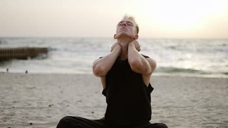 A-young-man-stretches-his-neck-while-doing-yoga-during-sunrise-on-the-beach.-Outdoor-sports-exercises