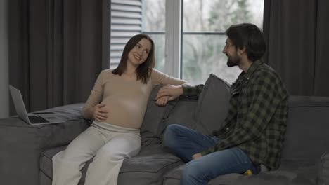 Woman-touching-pregnant-belly,-sitting-with-friend-or-husband-on-sofa