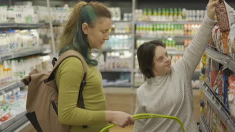 Down-syndrome-cheerful-girl-with-her-mother-taking-goods-from-shelf-in-supermarket,-side-view