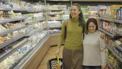 Lächelndes-Mädchen-Mit-Down-Syndrom-Und-Ihrer-Mutter-Im-Supermarkt,-Die-Mit-Einem-Einkaufskorb-Spazieren-Geht