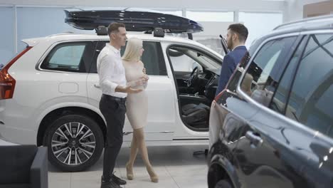Couple-talking-to-car-dealer-in-dealership-discussing-automobiles-looking-at-luxurious-new-white-model